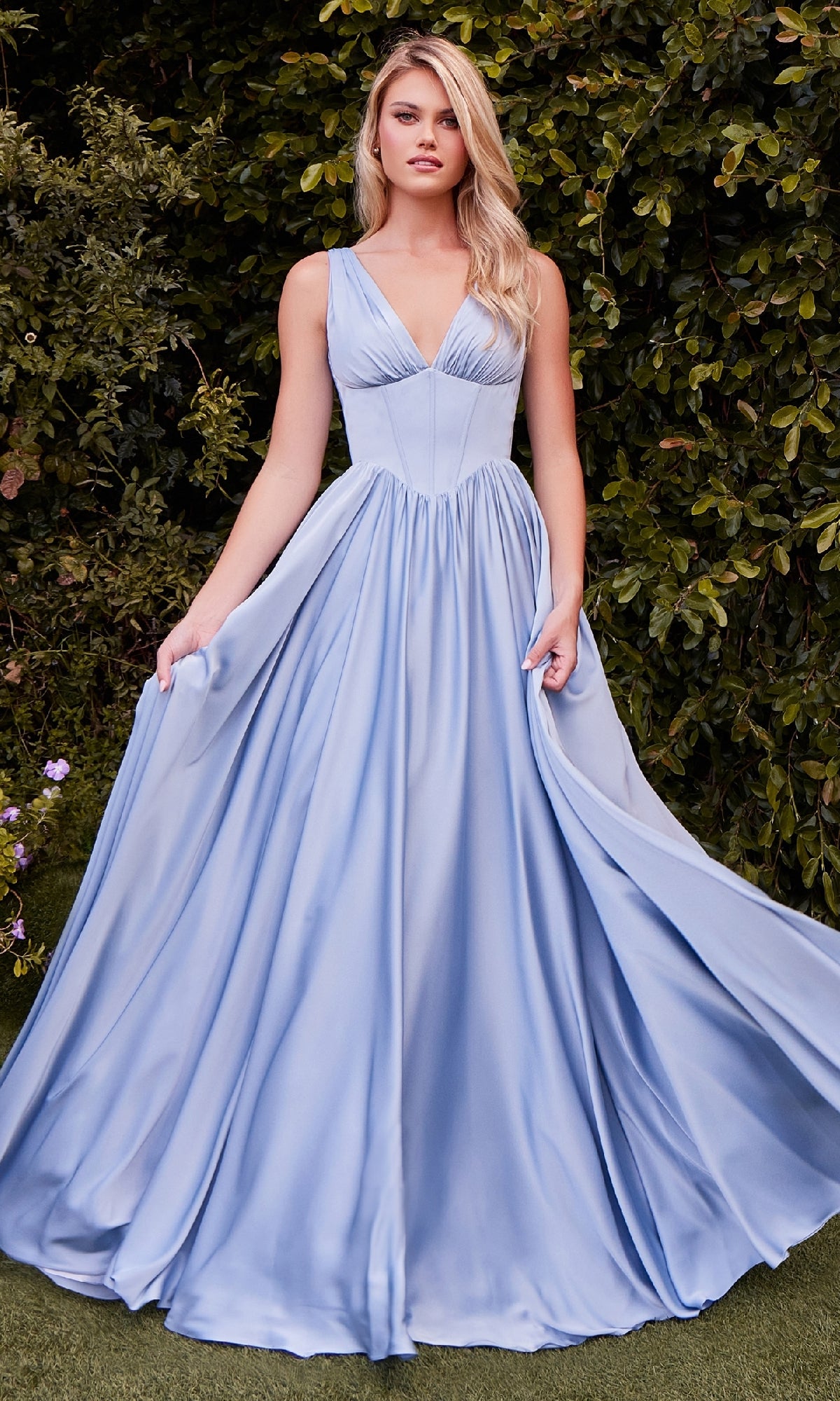 A woman in the Lace-Up Long A-Line Prom Dress: Ladivine 7447, a sleeveless light blue empire-waist corset gown, stands outdoors before lush green foliage, gracefully holding the flowing sides of her dress.